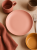 Widok z góry na matowy ceramiczny talerz w kolorze pastelowego różu, umieszczony pośrodku beżowego blatu. Aranżacja stołu w ciepłych odcieniach obejmuje kubek w kolorze terakoty, musztardową miseczkę, beżową czarkę, lnianą serwetkę oraz srebrny widelec.