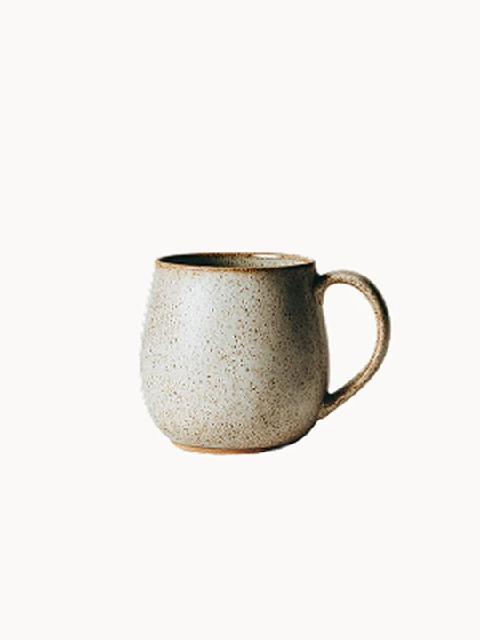 Nakrapiany, jasnobeżowy kubek ceramiczny o zaokrąglonym kształcie i solidnym uchu na białym tle. Powierzchnia pokryta jest drobnymi, brązowymi kropkami, odsłaniając naturalny, nieszkliwiony brzeg u samej podstawy.